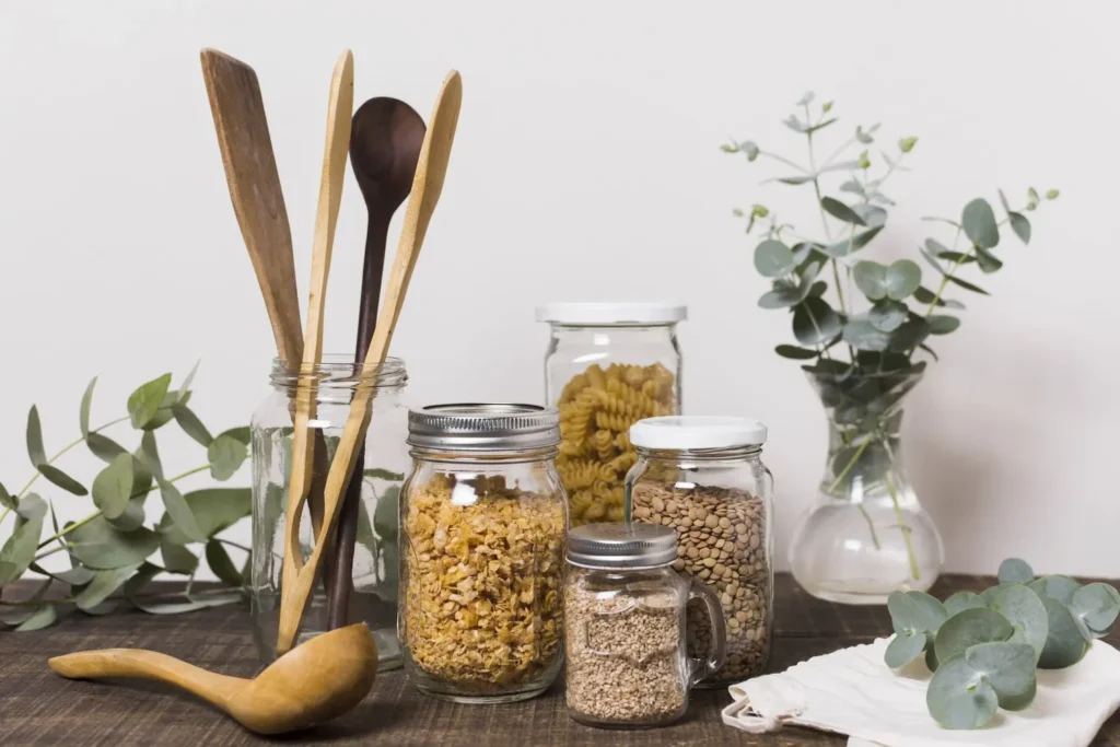 Bocaux en verre remplis de pâtes et de céréales, un bocal avec des ustensiles de cuisine écologiques, entourés de plantes vertes.